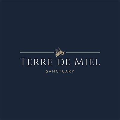 logo terre de miel représenté par un carré bleu foncé avec une bordure fine et dorée en son centre une abeille dorée sur lequel est incrit au centre terre de miel et blanc et sanctuary en doré / honey land logo represented by a dark blue square with a fine golden border in its center a golden bee on which is inscribed in the center honey land and white and sanctuary in gold 